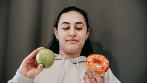 Content young female in hoodie deciding between sweet unhealthy doughnut and green ripe apple in light room